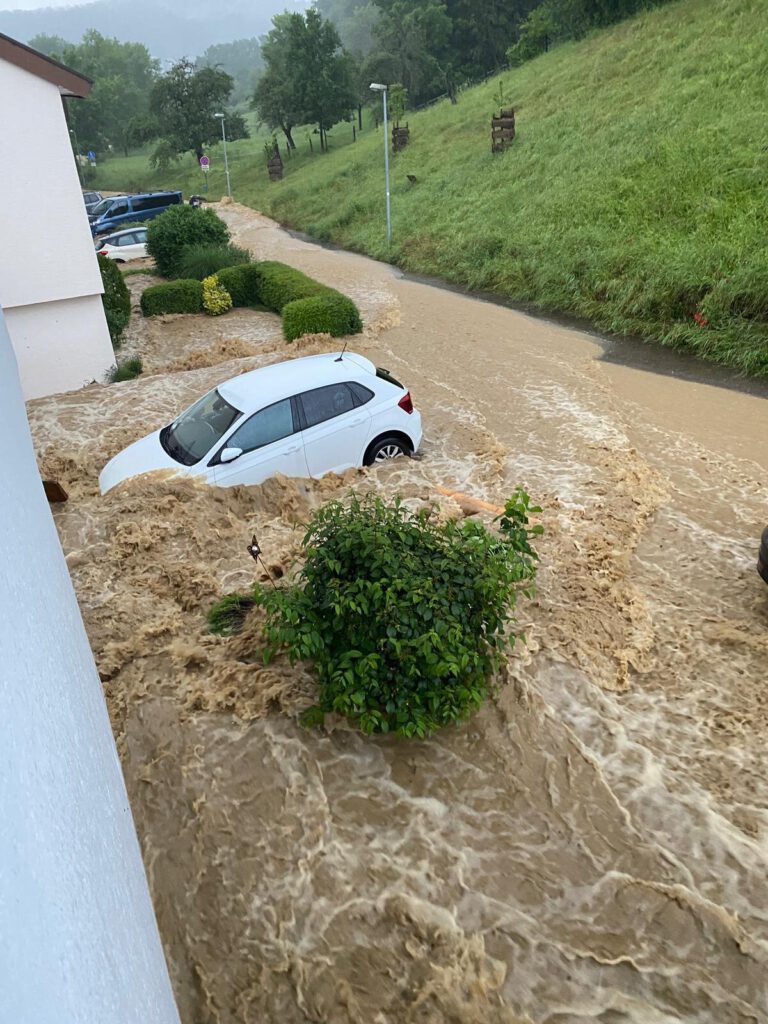 Braunes Hochwasser strömt durch eine Straße in Bünzwangen, ein Auto steht im Wasser